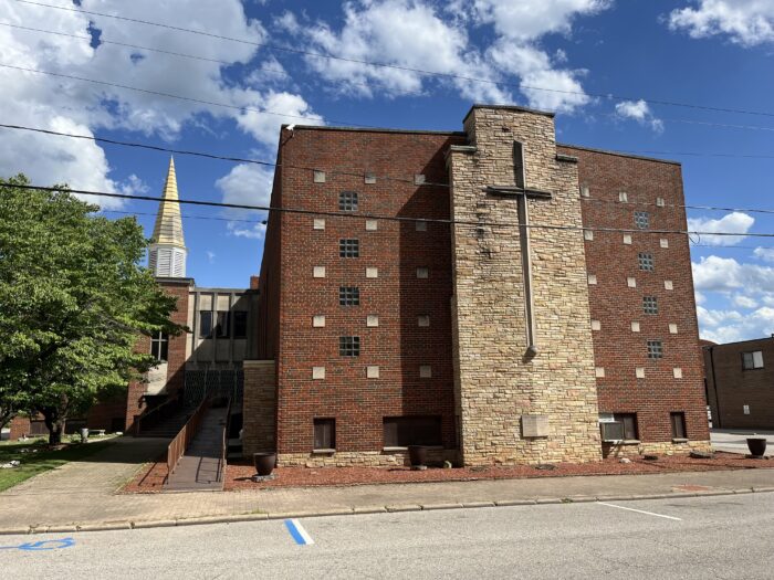 A tall brick church stands in an empty street. It displays a large cross on the side facing the camera. In the background, a church steeple rests against a clear blue sky.