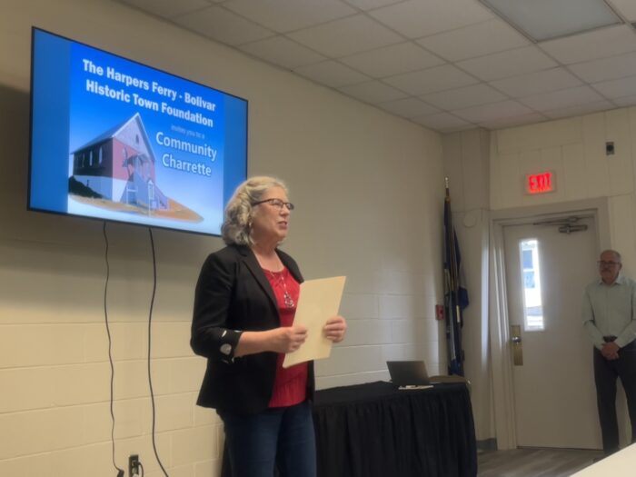 An older woman wearing a black jacket, jeans, dark pink shirt and glasses stands before a crowd that is off screen. Behind her is a TV monitor. She holds a sheet of paper and is speaking.
