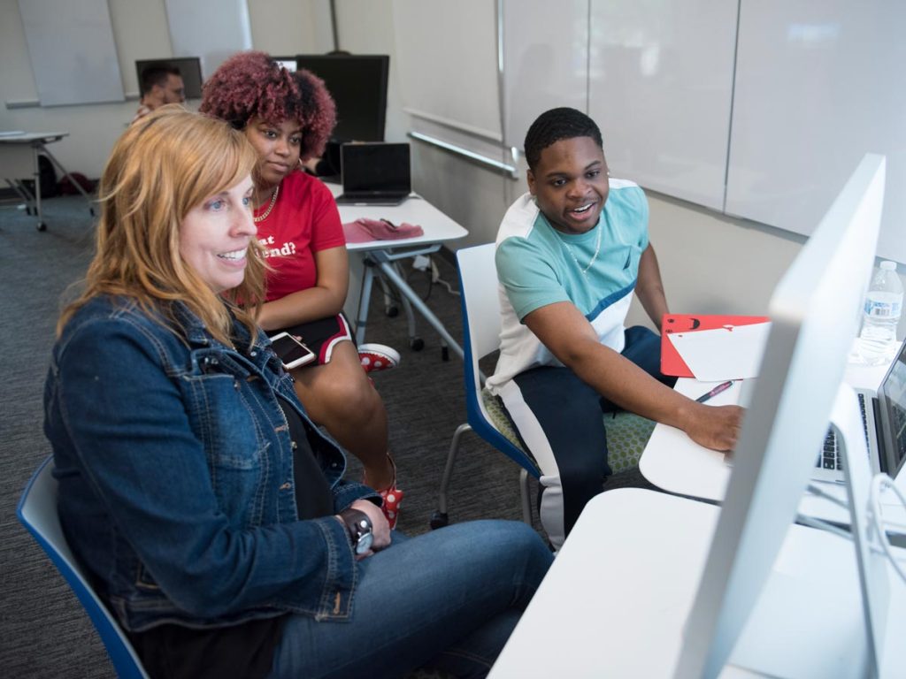 Three individuals in a modern classroom setting. A woman in a denim jacket is at the forefront, with two students behind her, one with curly red hair and the other working on a computer.