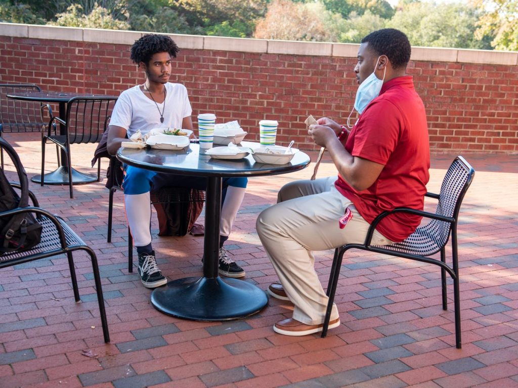 Two individuals sitting at an outdoor table on a brick patio, enjoying a meal. One person is wearing a mask.