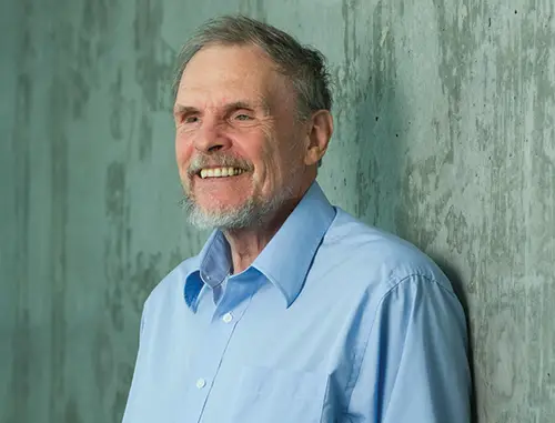 https://scienceline.org/2019/06/beyond-the-visible-what-shells-can-tell-us-about-our-world/ A close up of Geerat Vermeij wearing a light blue shirt with short grey hair and beard standing in front of a concrete wall