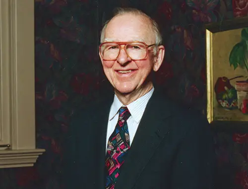 Edwin Krebs wearing large glasses, a black jacket and patterned blue and red tie standing in front of a patterned wallpaper