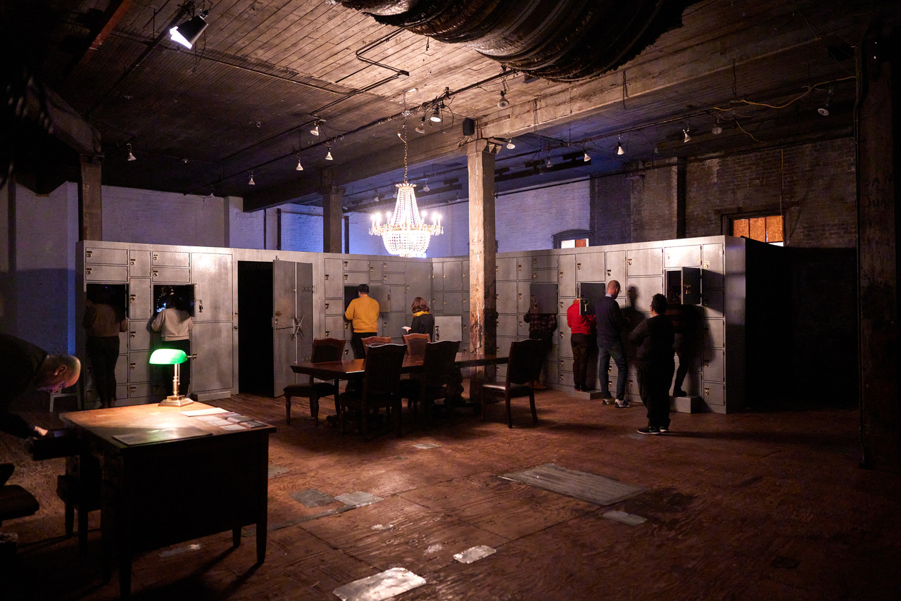 People gather around open and closed silver lockers in a foreboding warehouse-like space
