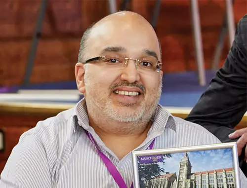 https://royalsociety.org/topics-policy/diversity-in-science/scientists-with-disabilities/hamied-haroon/ A close up of Hamied Haroon with short grey hair and facial stubble wearing glasses and a pale blue shirt holding a book and sat in a lecture theatre