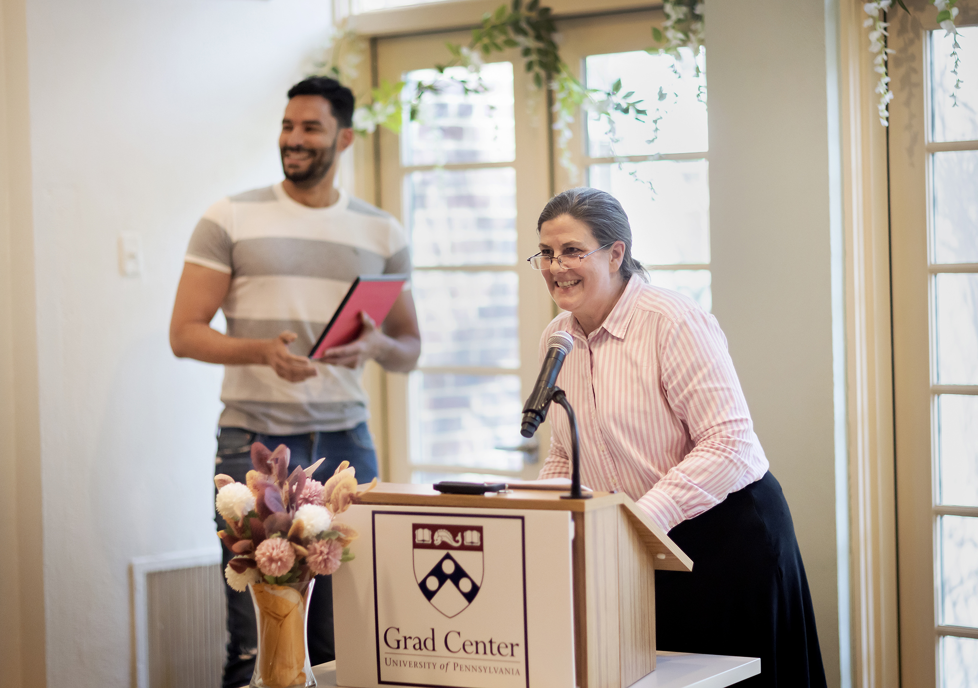 Speaker at a podium adorned with flowers, with a student off to the side holding a certificate.