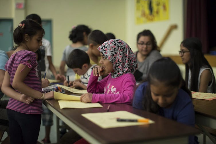 A child in a headscarf seen sitting at a desk in a classroom.