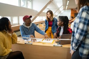 A diverse group of young adults converge around the desks to discuss their college work in a classroom at university.