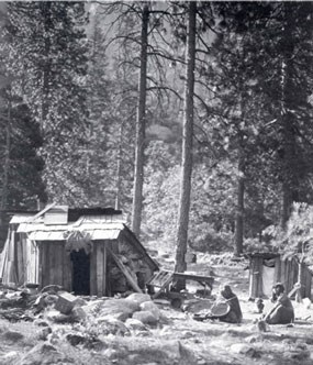 lt= Two American Indians sit on ground by their simple housing