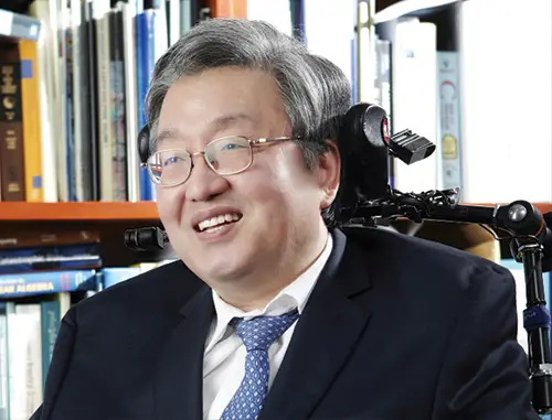 https://royalsociety.org/topics-policy/diversity-in-science/scientists-with-disabilities/sang-mook-lee/ A close up of Lee Sang Mook in a wheelchair with short grey hair wearing a black suit, white shirt and blue tie and glasses in front of shelves of books