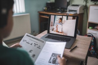 A student holds a workbook while watching a virtual lesson on a laptop computer.