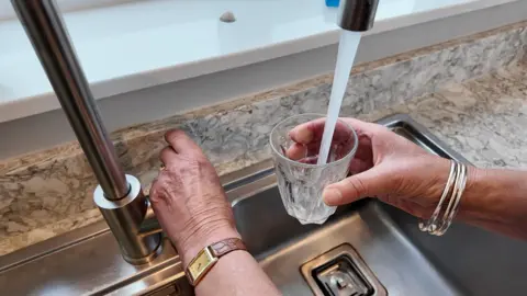 A view of a sink, with water flowing into a glass being held under the tap. Another hand is on the tap.