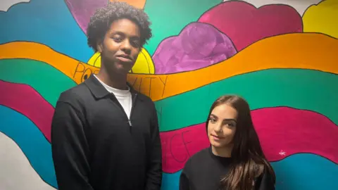 BBC Ridwan and Jayda. Ridwan has short, dark curly hair. He is wearing a white t-shirt and a black jumper. Jayda has long dark hair. She is wearing a black jumper. They are stood side by side in front of a colourful wall. They are looking at the camera and smiling.