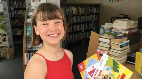 Corina Buckwell A young Wini is smiling in a red top, holding copies of comics at Ringmer Village Library in East Sussex.