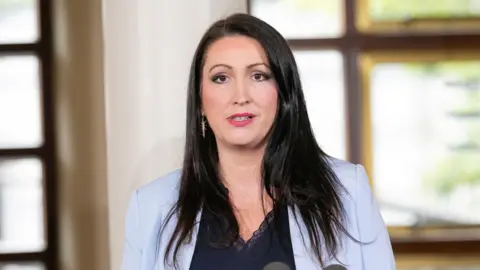 PA Emma is wearing a light blue blazer and navy vest top underneath.
She has long black hair and is wearing dangly gold earrings.