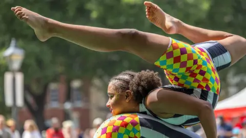 Paul Cox A young person in a colourful leotard doing a gymnastic trick on the back of someone else in the same outfit