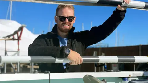Kyle Meredith Kyle Meredith is leaning against a boat. He wears a black fleece, black sunglasses and a black watch