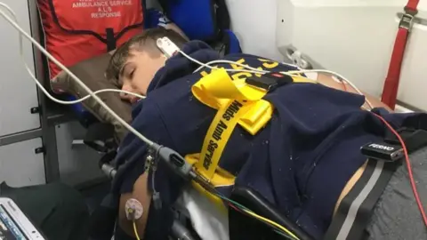 Xander Van der Poll A man lies face down on an ambulance stretcher, with a yellow securing belt around his back