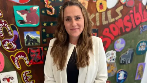 Alex Davies. She has long dark hair. She is wearing a white jacket and a black top. She is looking directly at the camera and smiling.