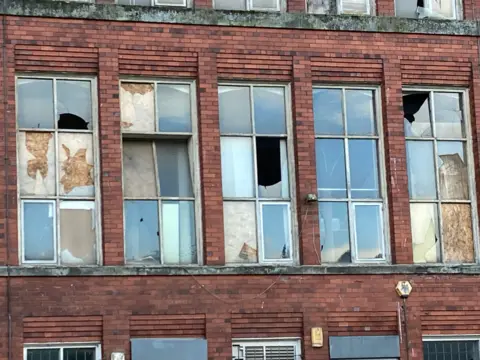 The picture shows windows in the Belper Mills buildings. A number of panes of glass are broken. In some areas, sheets of wood have been placed behind the broken glass. The image is a good example of how the old mill has fallen into disrepair.