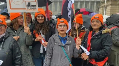 BBC A group of women are wearing orange hats with the GMB Union logo on them. They are holding banners and flags that call for equal pay. They are wearing coats and jackets, and people in the crowd behind them are holding umbrellas.