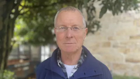 BBC Chris Weatherley stands outside, with hedges and a stone building in the background. He is wearing glasses and a blue jumper.