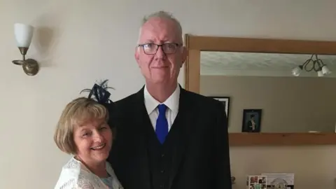 British Red Cross A smiling Jayne Radley (left) wears a black fascinator and pale blue-and-white dress and white patterned scarf. Next to her, her husband Steve wears a black suit, white shirt and dark blue tie.