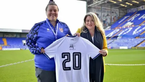 Tranmere Rovers FC Hannah Naylor wears a Tranmere top and dark trousers, while Jessica Gallier Booth stands next to her at the Tranmere ground. Ms Gallier Booth is wearing a mustard-coloured jacket, white top and black scarf. They are holding a Tranmere Rovers women's team shirt with the number 20 and The Martin Gallier Project logo on the back.