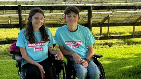 Molly Everitt Molly and her brother Stan, in wheelchairs, with a background of a field and a lake.