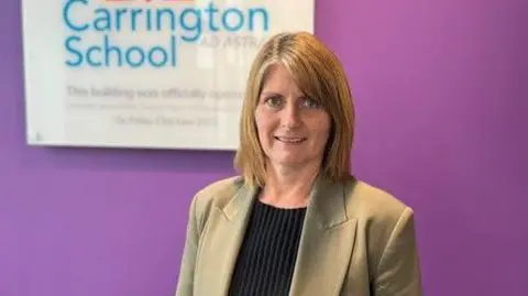Kerry Oakley in the corridor of her school near Redhill in Surrey. She is standing in front of a purple wall with a sign saying Carrington School behind her