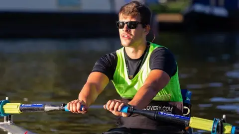 Nick B Images Xander Van der Poll rowing in a river, wearing a black shirt, green sports bib, and black sunglasses