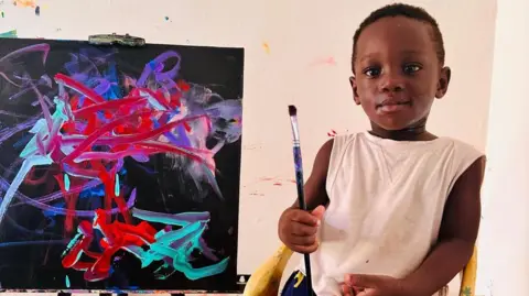 Chantelle Kuukua Eghan A toddler holding a paintbrush in front of a painting