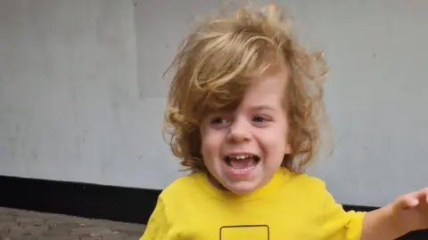 Contributed A photo of Wilfred. He is smiling and looking away from the camera. He has long, curly blonde hair and is wearing a yellow T-shirt. He is holding one arm away from his body.