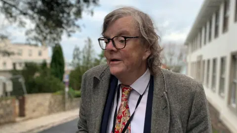 BBC Charles Parkinson standing in the street. He is wearing a grey jacket, a blue suit jacket, a white shirt and a patterned red, white and yellow tie. He has medium length curly hair and thick rimmed black glasses. The street in the background is blurred.