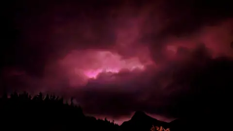 Muddy-Paws/BBC Weather Watchers Aurora appears pink in colour behind clouds and a hill.