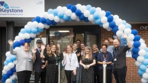 Oxford Health NHS Foundation Trust A group of people gathered together waving at the camera while stood under a blue and white balloon arch outside the front of a building
