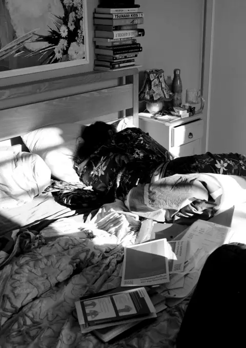Cora Slack A black-and-white photo of a person lying in bed surrounded by possessions