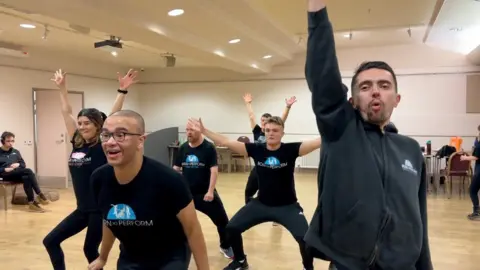 Carroll Weston/BBC Several men in black t-shirts and shorts dancing