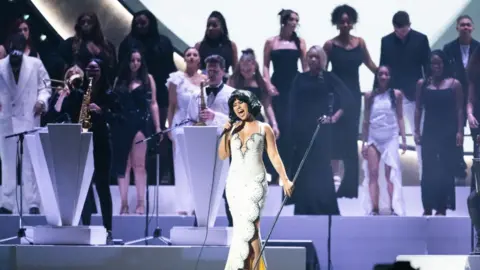 Getty Images Raye performing at the 2024 Brits with choir Flame Collective behind her. Raye has a short dark bob and wears a long white dress with black crystal detailing and scalloped edges. Behind her are musicians with brass instruments as well as the singers from the Flame collective choir. The other musicians are all dressed in black or white