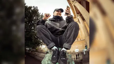 Ivaylo Erev Danny Pierce sitting on top of a fountain statue and smiling at his phone