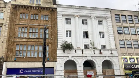 The Corn Exchange, a white Grade II listed building. At the front of the building are three archways, which are partially boarded up. The building has a Cancer Research on one side and a casino on the other.