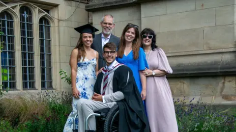 PA Media/University of Bristol Xander in his graduation robes, sat in a wheelchair, with his wife and three family members around him, smiling at the camera, outside a University of Bristol building
