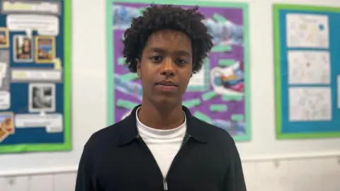 Ridwan. He has short, dark curly hair. He is wearing a white t-shirt and a black jumper. He is looking at the camera and smiling.