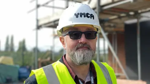 BBC A man with a beard looking directly into the camera. He has a grey beard and dark glasses and is wearing a high vis jacket and hard hat. He is standing in front of a construction site covered in scaffolding.