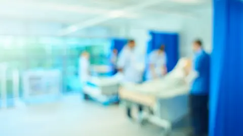 Sturti/Getty Creative A senior female nursing sister demonstrates the the various equipment on the training ward whilst a male staff nurse shows the medical mannequin to another group of medical student nurses. They are all standing around the hospital beds. The whole scene is out of focus.
