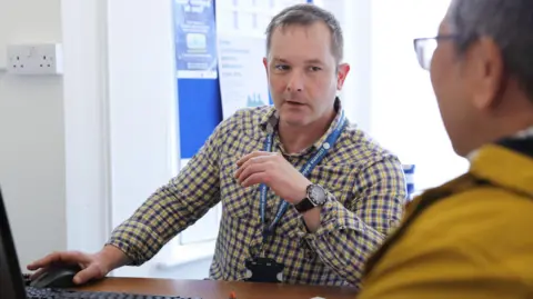 Citizens Advice Cornwall A male Citizens Advice volunteer wearing a blue and yellow chequered shirt sat at a desk with a computer on it talks to a person sat opposite him.