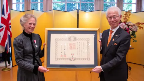 Beatrice McCosh  Jane Hasell-McCosh on one side, with a medal on her jacket, holding a framed certificate in Japanese, with Hajime Hayashi on the other side wearing a suit and orange tie, also holding the frame.