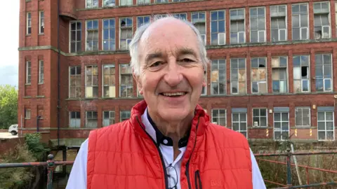 BBC The picture is Derek Latham who is the chair of the Derbyshire Historic Buildings Trust. He is smiling and wearing a red, sleeveless coat and light blue shirt. Behind him are the old Belper Mills buildings