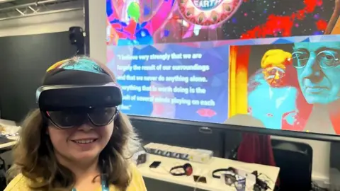 University of Gloucestershire Beth Hands, a young female university student, wearing a VR headset as she stands in front of a colourful screen bearing an image of Gustav Holst