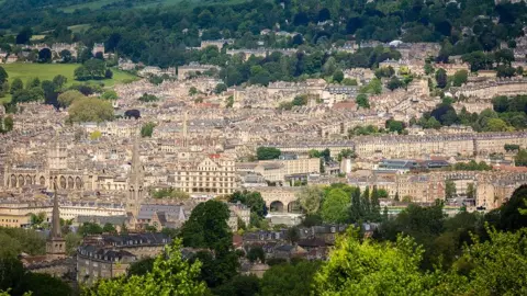 University of Bath The city of Bath as captured from high up on a hill. You can see lots of ornate historic buildings interspersed with trees and green fields.