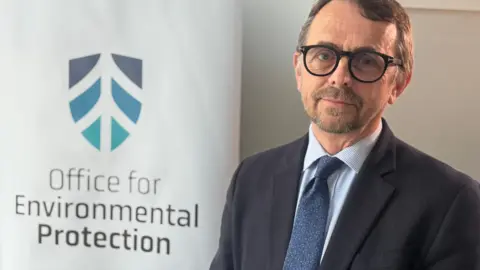 Robbie McDonald, wearing a dark suit jacket, blue shirt, dark blue tie and black-rimmed glasses, looks towards the camera. He is standing in front of a branded sign which says "Office for Environmental Protection."